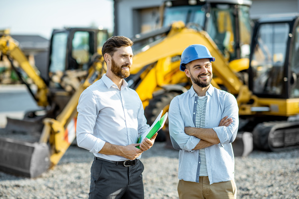 Builder choosing heavy machinery for construction with a sales consultant standing with some documents on the open ground of a industrial equipment vendor shop