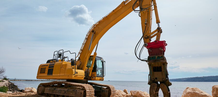 Yellow tracked excavator with hydraulic claw attachment working on a rocky coastal construction site