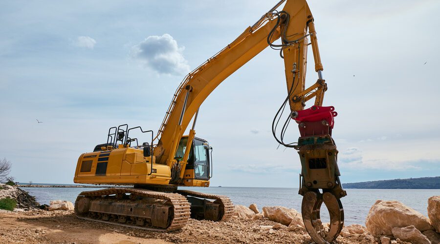 Yellow tracked excavator with hydraulic claw attachment working on a rocky coastal construction site