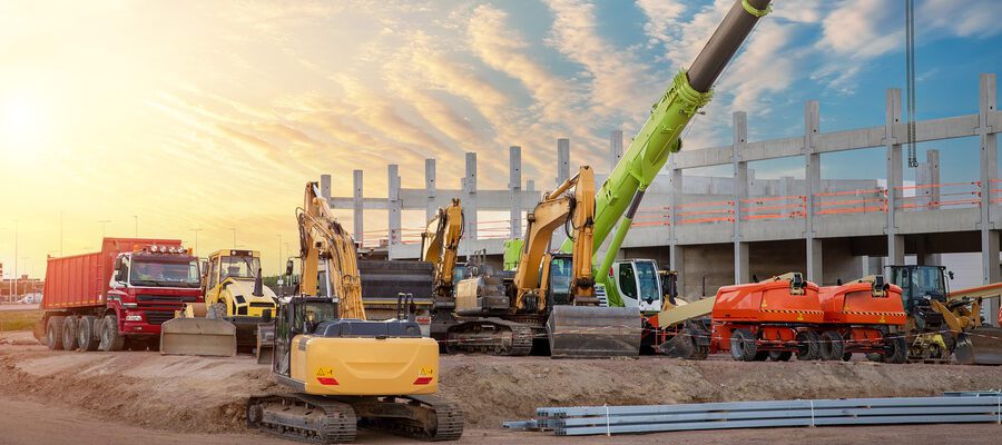 Many different multiclored colorful heavy industrial machinery equipment at construction site parking area against warehouse building city infrastructure development. Commercial vehicles rental sale