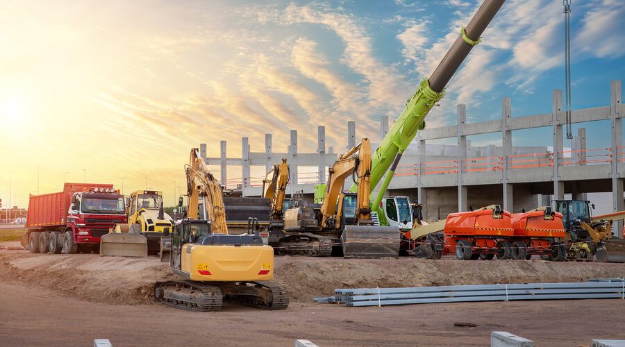 Many different multiclored colorful heavy industrial machinery equipment at construction site parking area against warehouse building city infrastructure development. Commercial vehicles rental sale