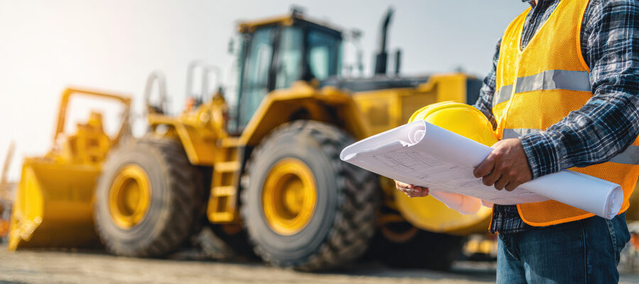 An engineer stands near a large yellow construction vehicle, holding a rolled blueprint for mantenance of the heavy equipments