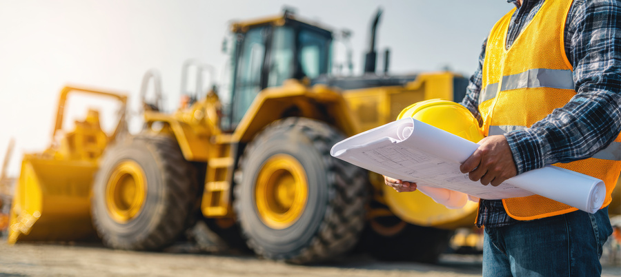 An engineer stands near a large yellow construction vehicle, holding a rolled blueprint for mantenance of the heavy equipments