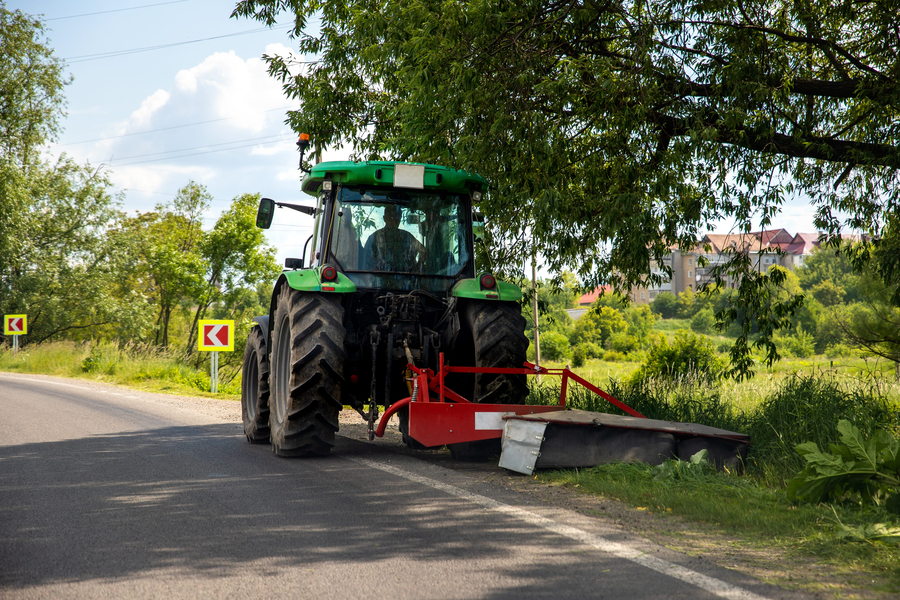 Big modern industrial tractor machine cutting green grass with mowing equipment along country roadside